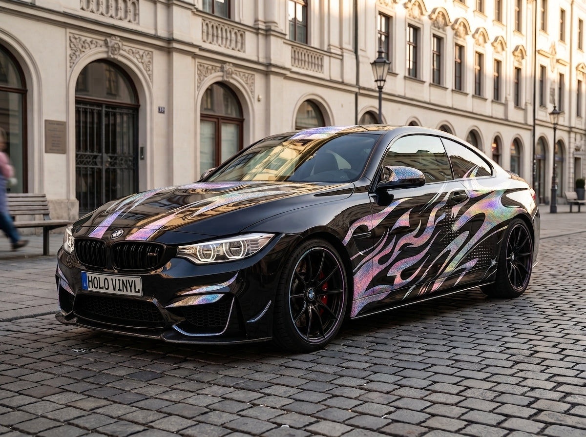 A black BMW M4 coupe parked on a cobblestone street in front of a historic building, featuring a holographic iridescent vinyl wrap with black flame graphics and white website text on the side.