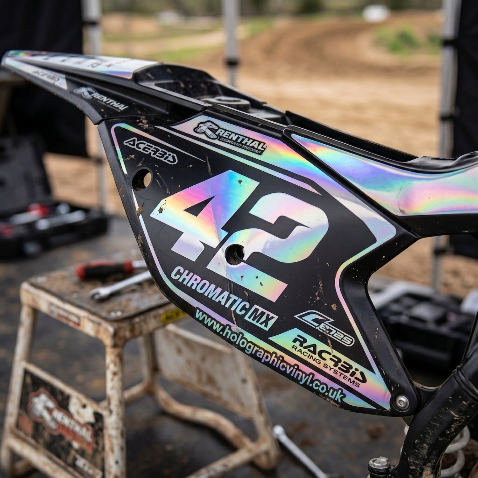 A close-up of a motocross bike body panel with a large holographic '42' decal, showing an iridescent rainbow effect on a black background. Visible decals include 'Chromatic MX', 'Rentalh', 'AG&FOLD', and 'RACERS'.